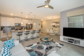Una cocina moderna con una isla central, taburetes de bar, un comedor con mesa y sillas, una sala de estar con sofá y mesa de centro, y un televisor de pantalla plana montado sobre un soporte.