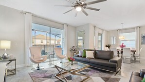 A modern living room with a glass coffee table, a sofa, and a chair. There is a ceiling fan, a dining area with a table and chairs, and large windows with curtains.