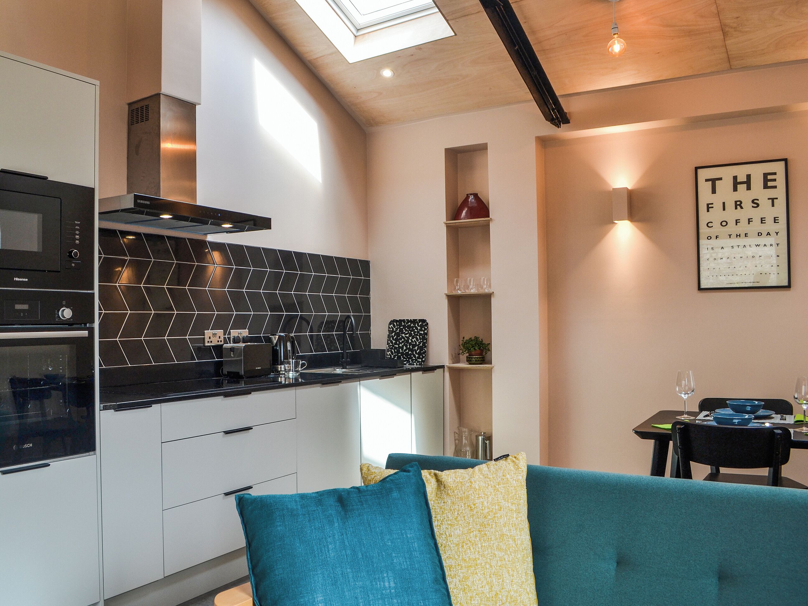 A modern kitchen with a black herringbone backsplash, a built-in oven, and a coffee machine. There is a teal sofa with yellow pillows, a dining table with a wine glass, and a framed quote on the wall.