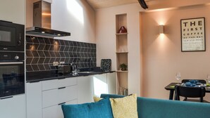 A modern kitchen with a black herringbone backsplash, a built-in oven, and a coffee machine. There is a teal sofa with yellow pillows, a dining table with a wine glass, and a framed quote on the wall.