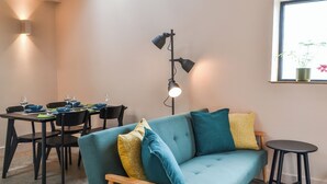 A modern living room with a teal sofa, wooden armchair, dining table with chairs, and a floor lamp.