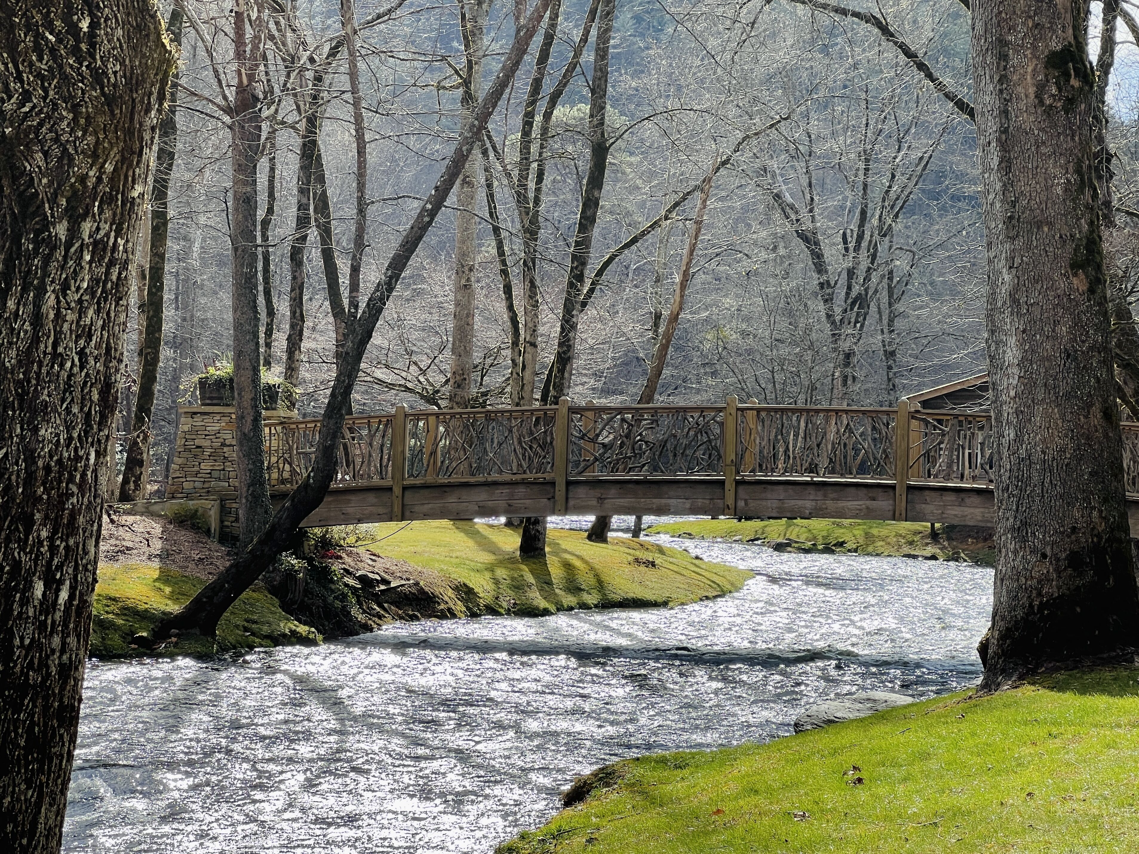 Un pont en bois enjambant une rivière, avec un mur de pierre et une zone herbeuse d’un côté.