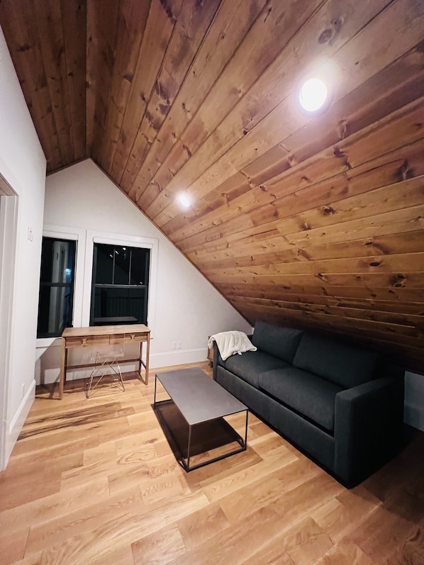 A loft-style room with a sloped ceiling, wooden beams, a dark gray sofa, a wooden desk, and a bench.