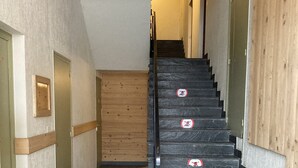 Una escalera con escalones negros y barandilla de madera, que lleva a una habitación con puerta de madera y un cuadro enmarcado en la pared.