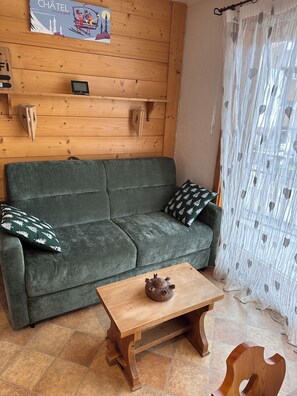 A cozy room with a wooden wall, a green sofa, a small wooden table, and a window with sheer curtains. - Apartment in the center of the Portes du Soleil village (Châtel)
