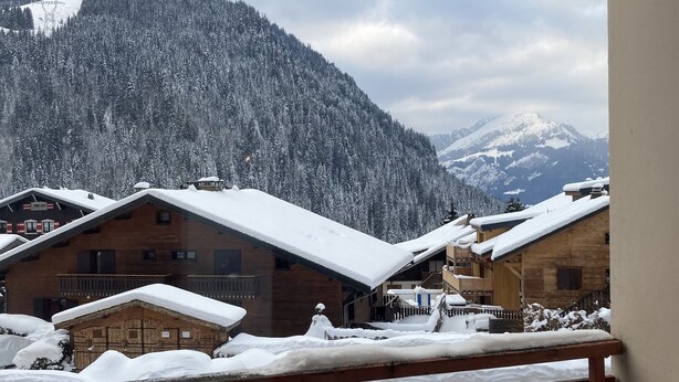Vista de un pueblo de montaña con edificios de madera cubiertos de nieve, un coche verde estacionado al frente y una valla de madera.