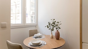 Een eetgedeelte met een ronde houten tafel voor twee personen, twee stoelen, een vaas met groen loof en een raam met uitzicht op gebouwen.