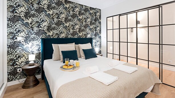 Un lit bien fait, avec une tête de lit bleue, une table de chevet sur laquelle se trouve une lampe, et un plateau de petit-déjeuner avec des croissants et du jus d'orange.