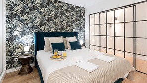 Un lit bien fait, avec une tête de lit bleue, une table de chevet sur laquelle se trouve une lampe, et un plateau de petit-déjeuner avec des croissants et du jus d'orange.