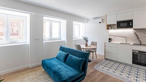 Un appartement moderne avec un canapé bleu, un coin repas et une cuisine dotée de meubles blancs et d’appareils électroménagers.