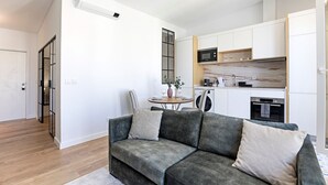 Un espacio habitable compacto con sofá gris, una pequeña mesa de comedor y una zona de cocina con armarios blancos y un microondas.