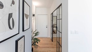 Un pasillo con obras de arte abstracto enmarcadas, una planta en maceta y una cama visible a través de una puerta de vidrio.