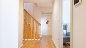 Ein Flur mit einer Holztreppe, die zu einer Tür führt, einem Stuhl mit Hunde-Zahn-Muster und einer weißen Wand mit einem Brandschutznachweis.