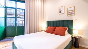 A hotel room with a bed, two bedside lamps, a window with curtains, and two framed pictures on the wall.