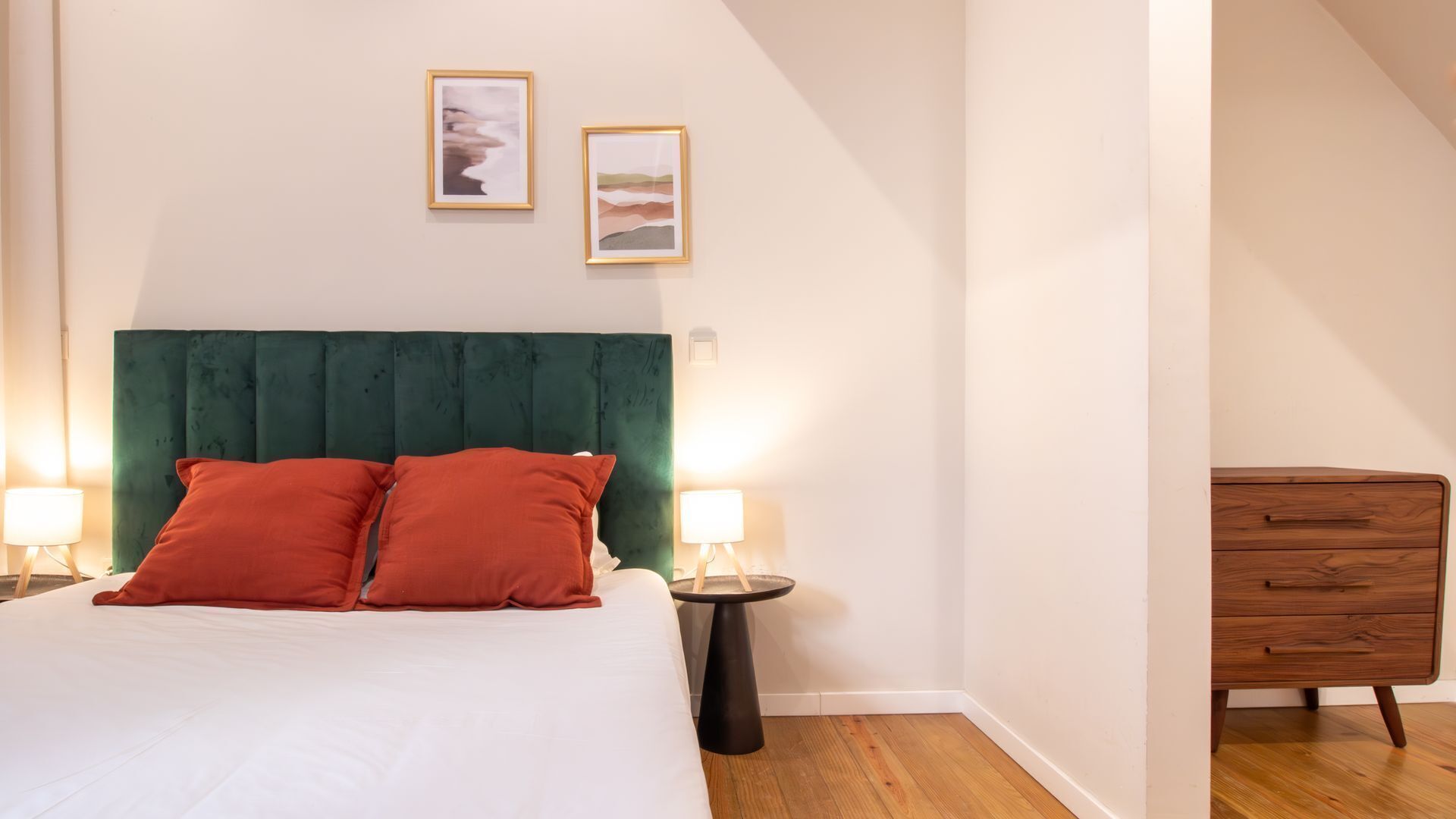 A bedroom with a bed, two pillows, a bedside table with a lamp, a dresser, and two framed pictures on the wall.