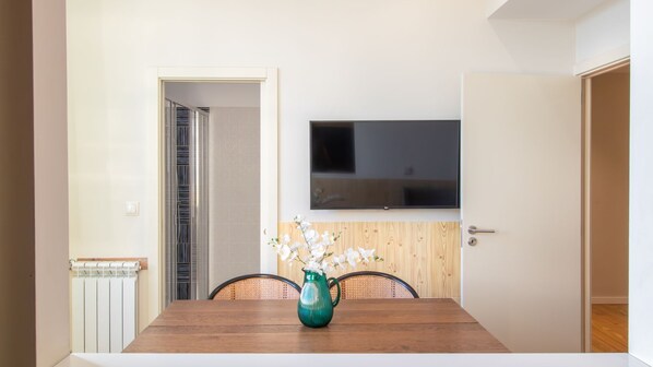 A dining area with a wooden table, chairs, a green vase with flowers, a TV mounted on the wall, and a door leading to another room.