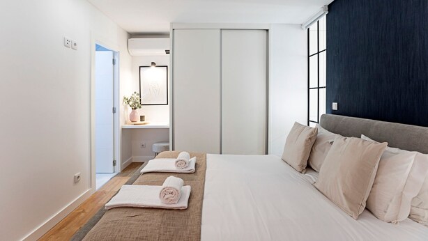 A modern hotel room with a large bed, a bedside table with a vase, and a view of the city through floor-to-ceiling windows. - Urban House - D - by LovelyStay (Porto)