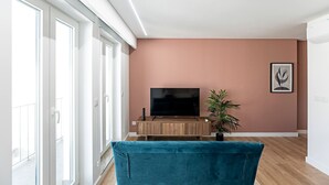 A modern living room with a large flat-screen TV on a wooden stand, a teal sofa, and a potted plant. - Urban House - D - by LovelyStay (Porto)