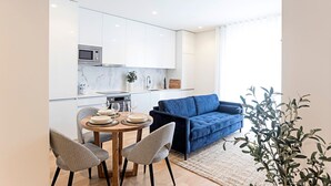 A modern kitchen with white cabinets, a microwave, and a sink. There is a blue sofa and two chairs in a dining area with a round table.