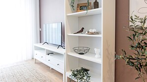 A modern living room with a flat-screen TV on a white TV stand, a white shelving unit with decorative items, and a potted plant.