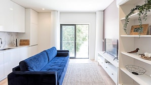 A modern living room with a blue sofa, a TV on a white stand, and a balcony with a view of greenery.
