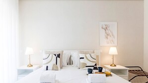 A hotel room with a neatly made bed, two bedside lamps, a framed artwork on the wall, and a small table with a tray of breakfast items.