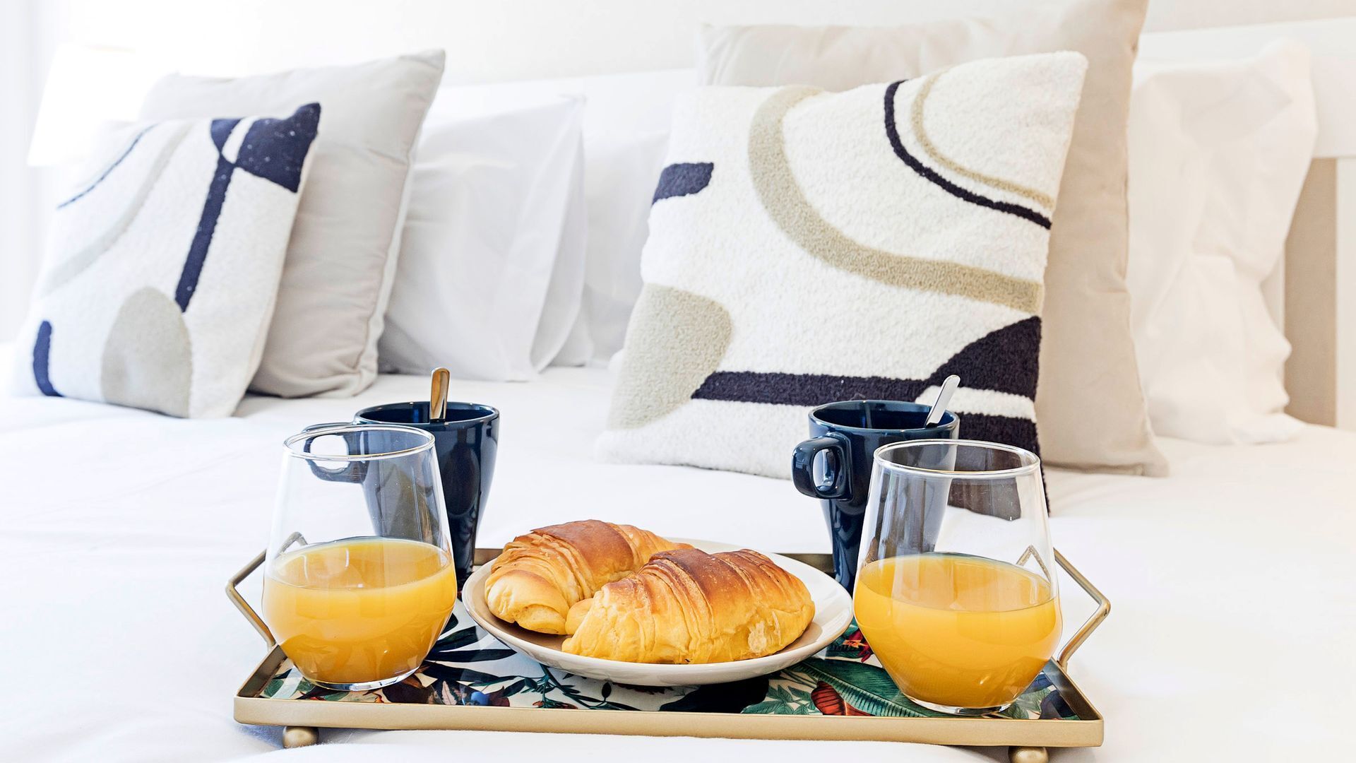 Bandeja de desayuno con croissants, zumo de naranja y café sobre una cama con ropa blanca y almohadas estampadas.