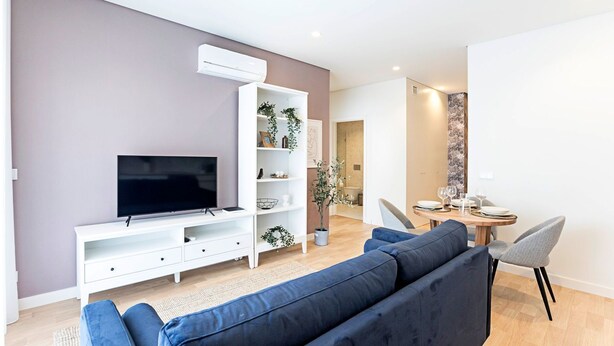 A modern living room with a flat-screen TV, a white entertainment unit, a blue sofa, and a dining area with a wooden table and chairs.