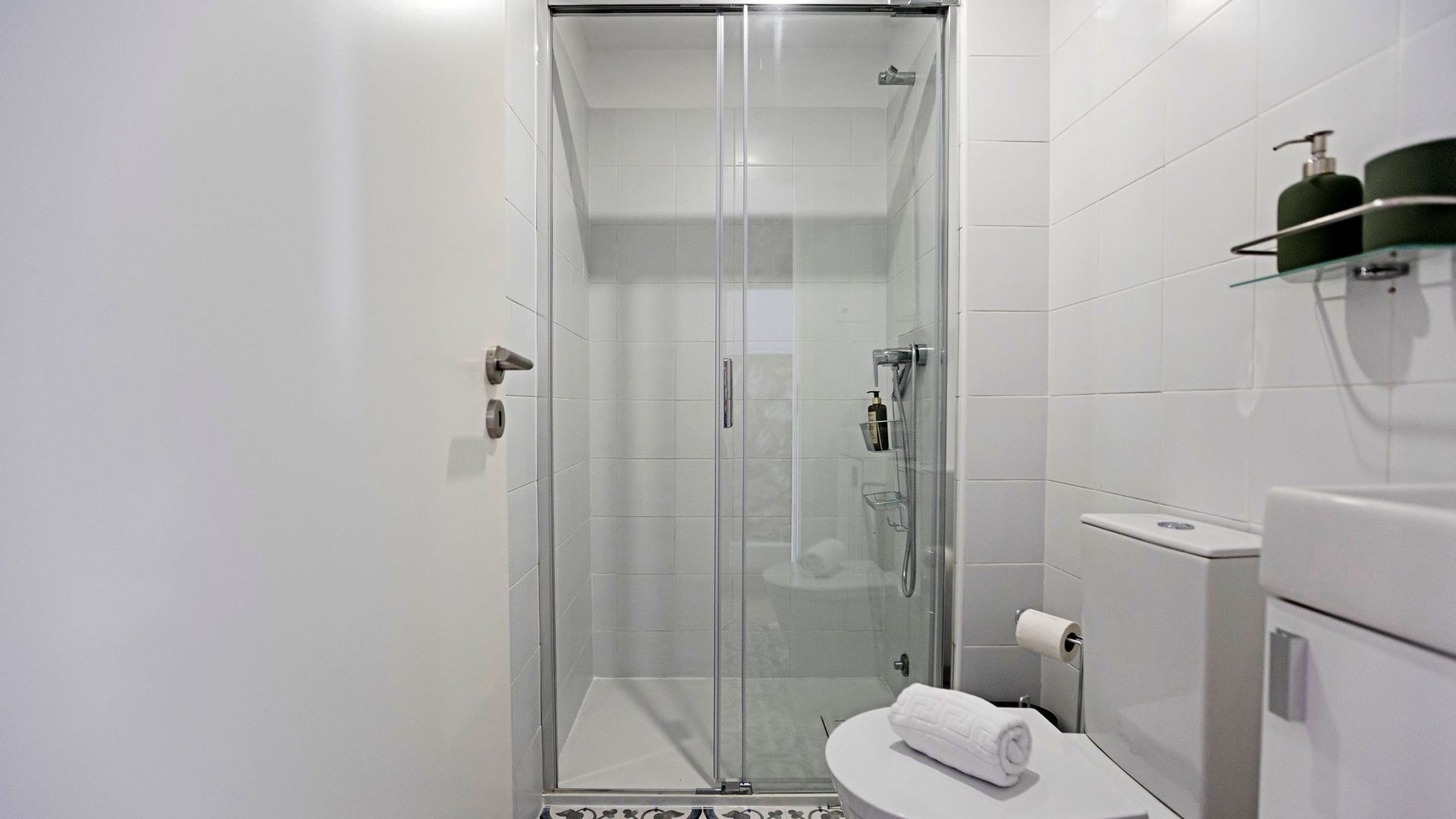 A modern bathroom with a glass-enclosed shower, white tiled walls, a toilet, and a towel on the sink.