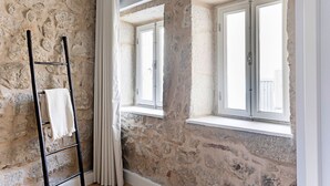 Um quarto com paredes de pedra, uma escada de metal preto e duas janelas com portadas brancas.