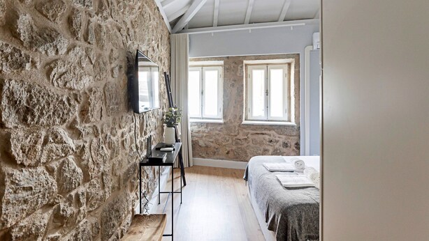 Quarto com paredes de pedra, uma cama, uma escrivaninha e duas janelas.