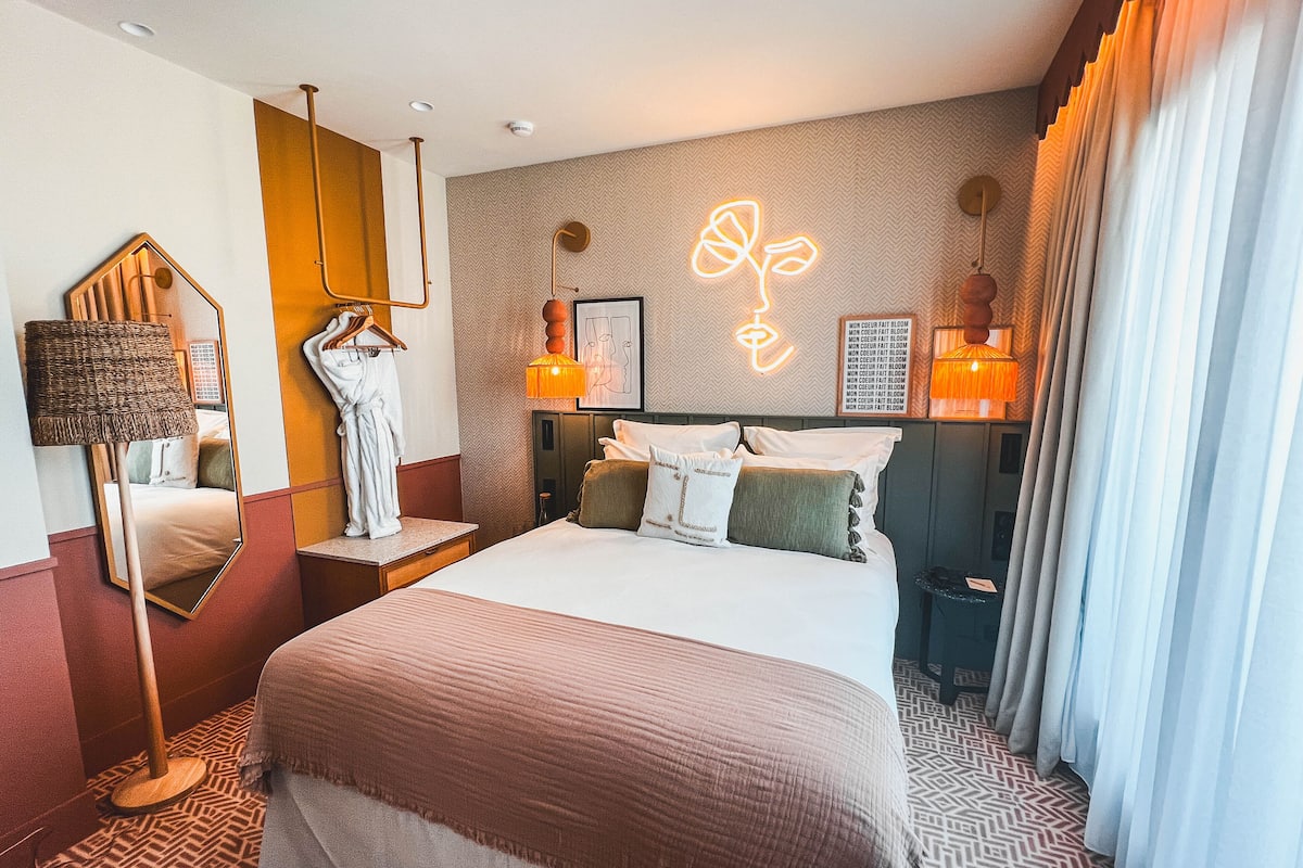 A hotel room with a large bed, a mirror, a hanging robe, a wall-mounted lamp, and a neon sign.