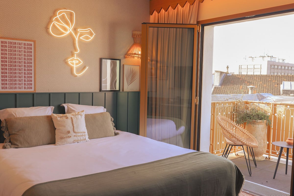 A hotel room with a bed, a neon flower decoration, a balcony with a chair and a potted plant.