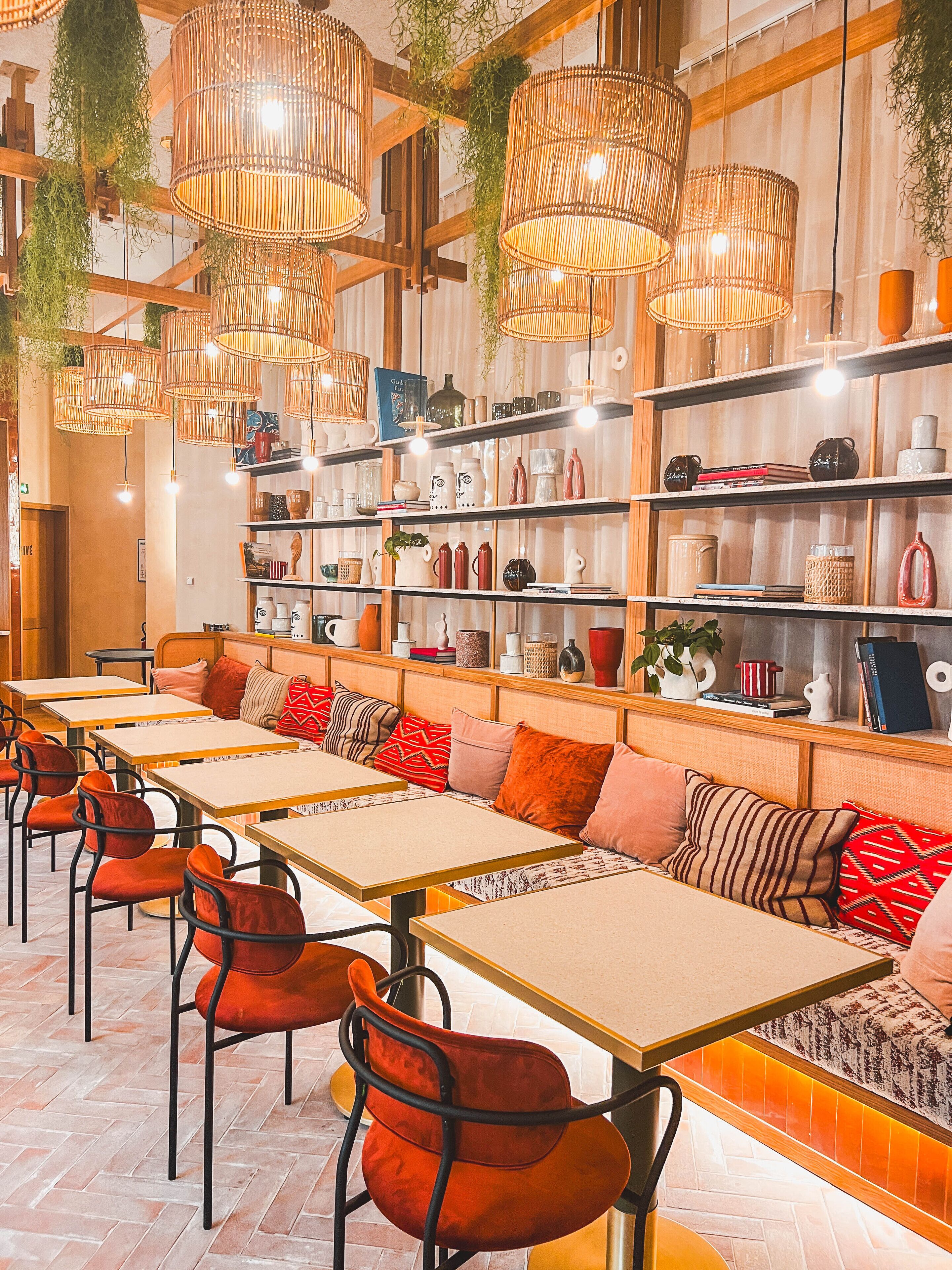 A restaurant interior with wooden tables and chairs, hanging wicker lamps, and shelves with various items.