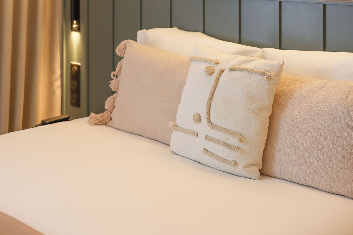 A neatly arranged bedroom with a bed, pillows, a wall-mounted light fixture, and a framed artwork.