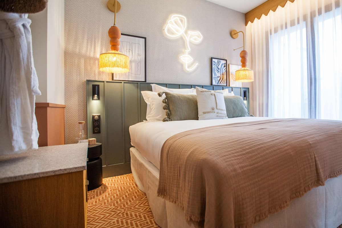 A hotel room with a large bed, two bedside lamps, a headboard with artwork, a nightstand, and a window with curtains.