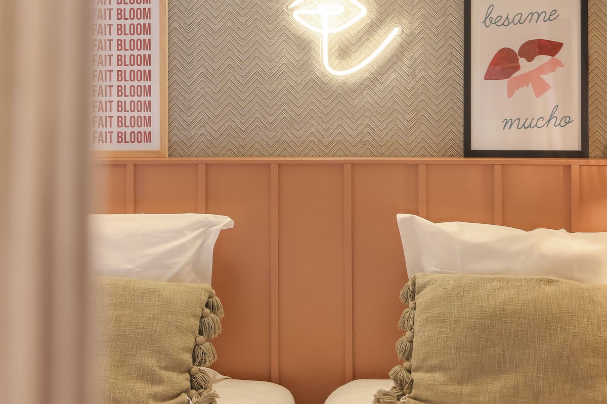 A hotel room with a herringbone-patterned wallpaper, a neon light art of a flower, a framed quote, and two beds with beige pillows.
