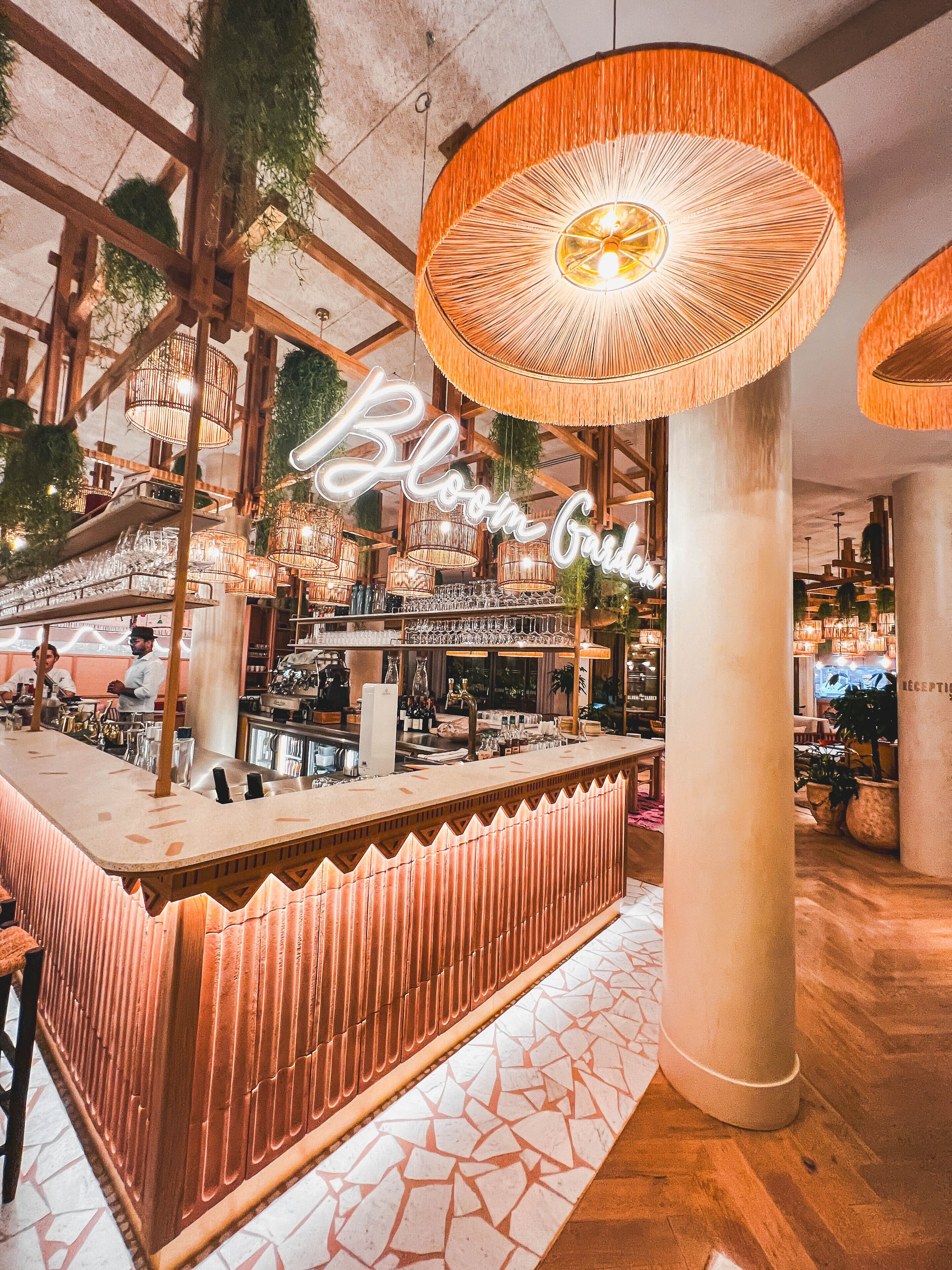 A bar area with a wooden counter, hanging wicker lamps, and a neon sign that reads "Bloom Garden".