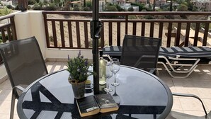 Una mesa redonda de cristal con sillas, una planta en maceta, un libro y dos copas de vino en un balcón con vistas a un pueblo costero.