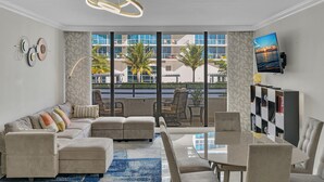 A modern living room with a sectional sofa, a dining table, and chairs. There is a large window with a view of palm trees and buildings.