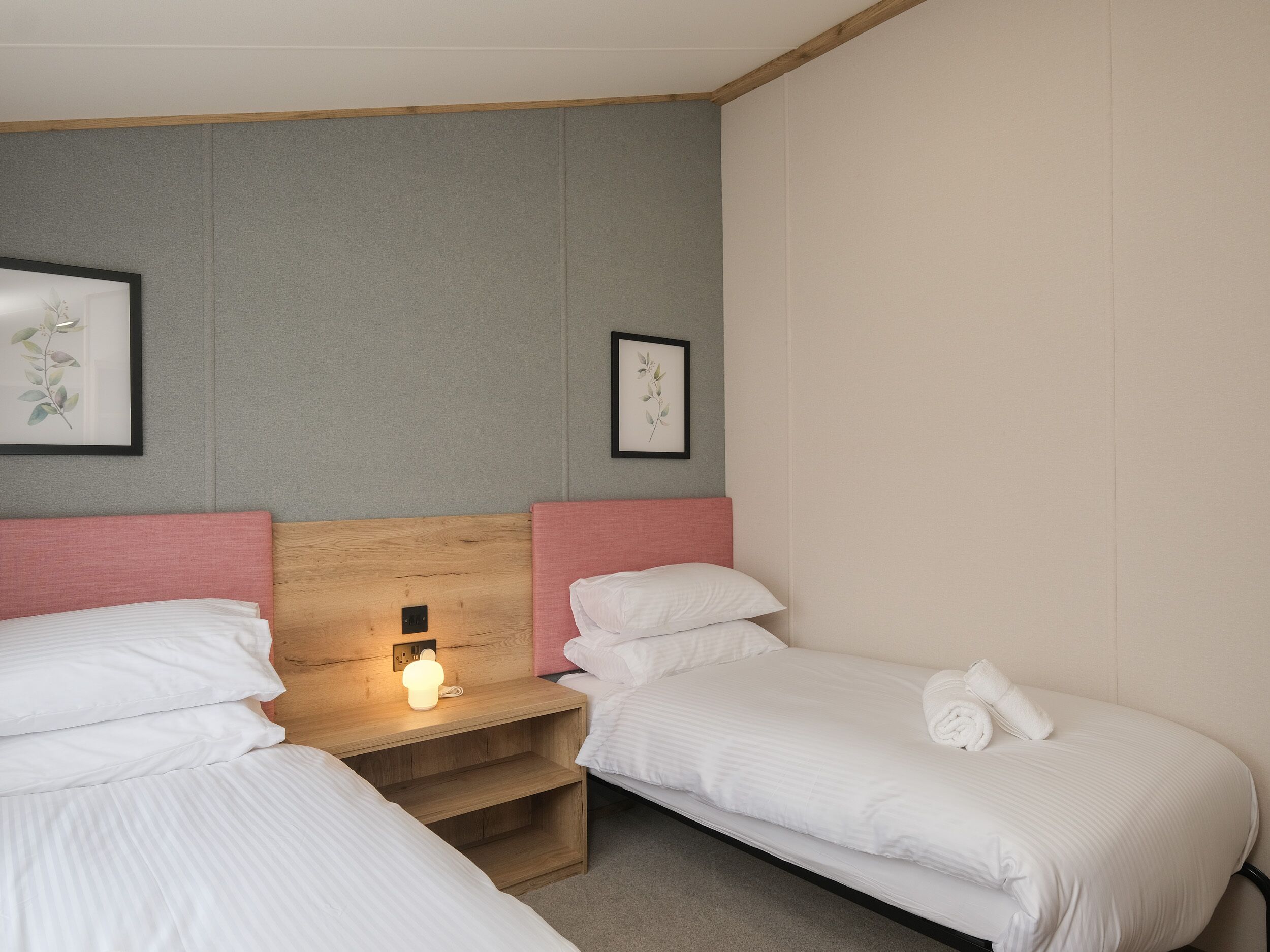 Una habitación de hotel con dos camas individuales, una mesita de noche de madera con una lámpara, y dos impresiones botánicas enmarcadas en la pared.