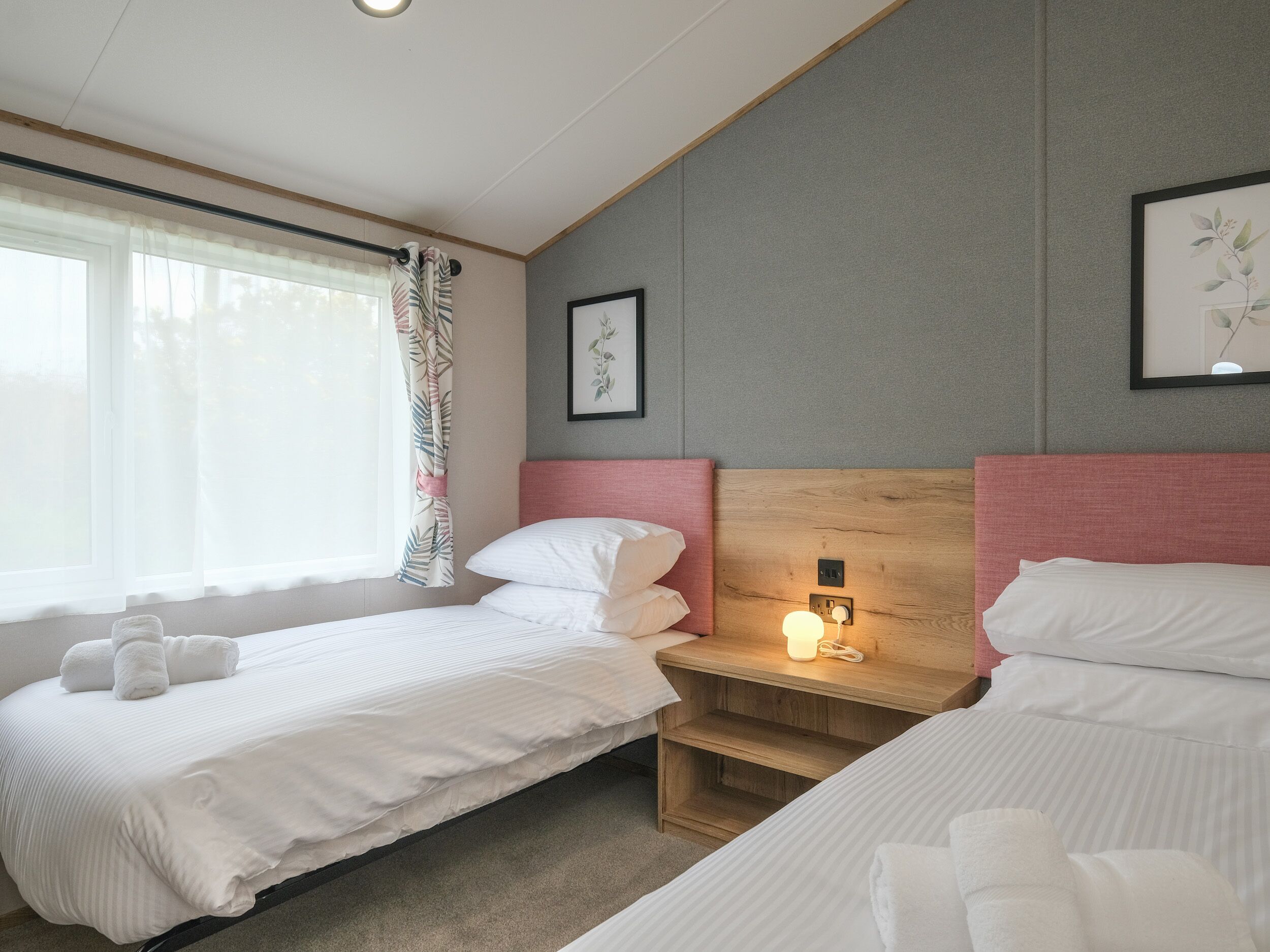 A hotel room with two beds, a wooden headboard, a bedside table with a lamp, and a window with sheer curtains.