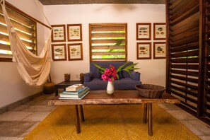 A living room with a hammock, a sofa, a coffee table with books and a vase of flowers, and framed fish illustrations on the wall. - CAMP BAY LODGE (Roatan)