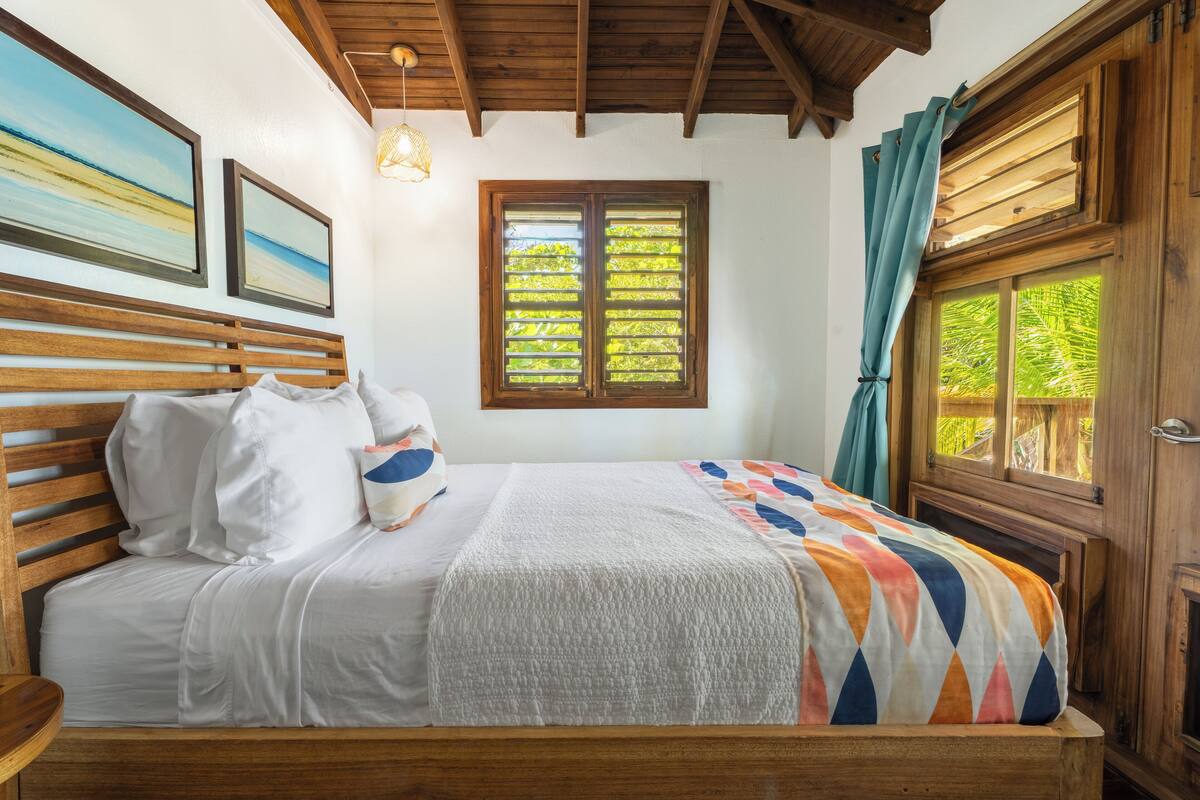 A bedroom with a wooden bed, white bedding, a colorful blanket, a window with shutters, and a view of greenery outside.