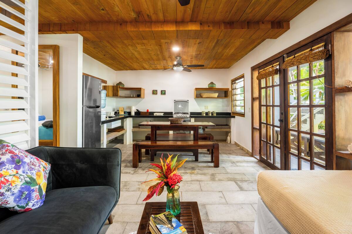 A spacious living area with a dark sofa, a coffee table with books and a vase, a wooden ceiling, and a view through large windows.