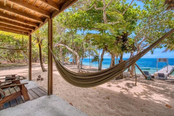 Una hamaca debajo de una estructura de madera en una playa con palmeras y vista al océano.