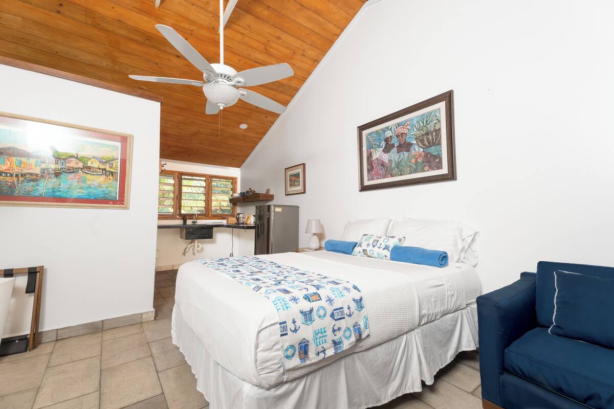 A bedroom with a bed, a blue armchair, a ceiling fan, and a painting on the wall.