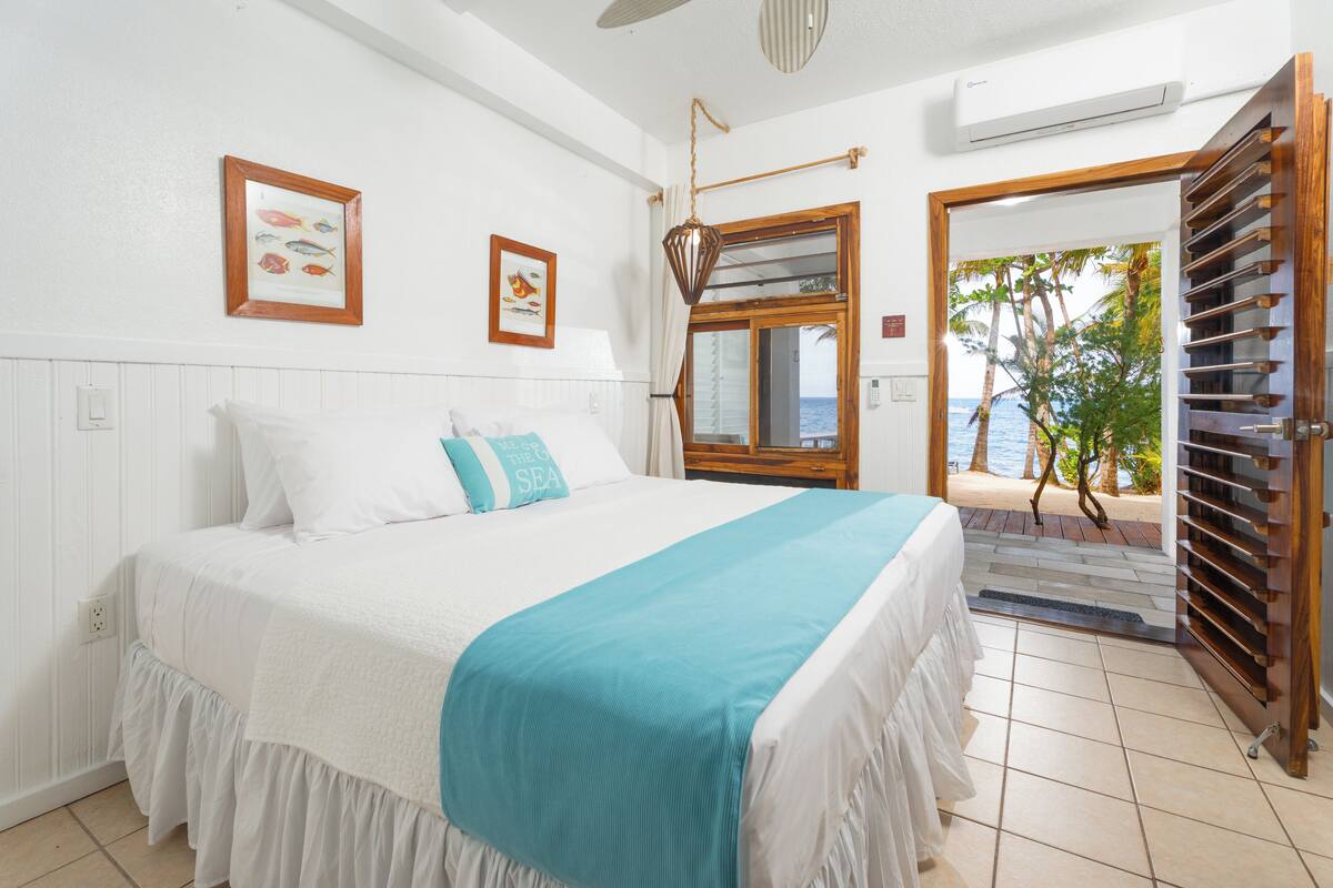 A bedroom with a bed, a window with a view of palm trees and the sea, and a wooden door leading to an outdoor area.