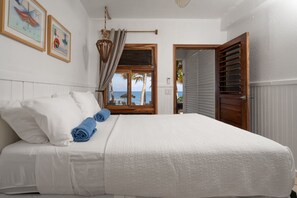 Habitación de hotel con cama, ventana con vista a palmeras y al mar, y puerta que da a un balcón.