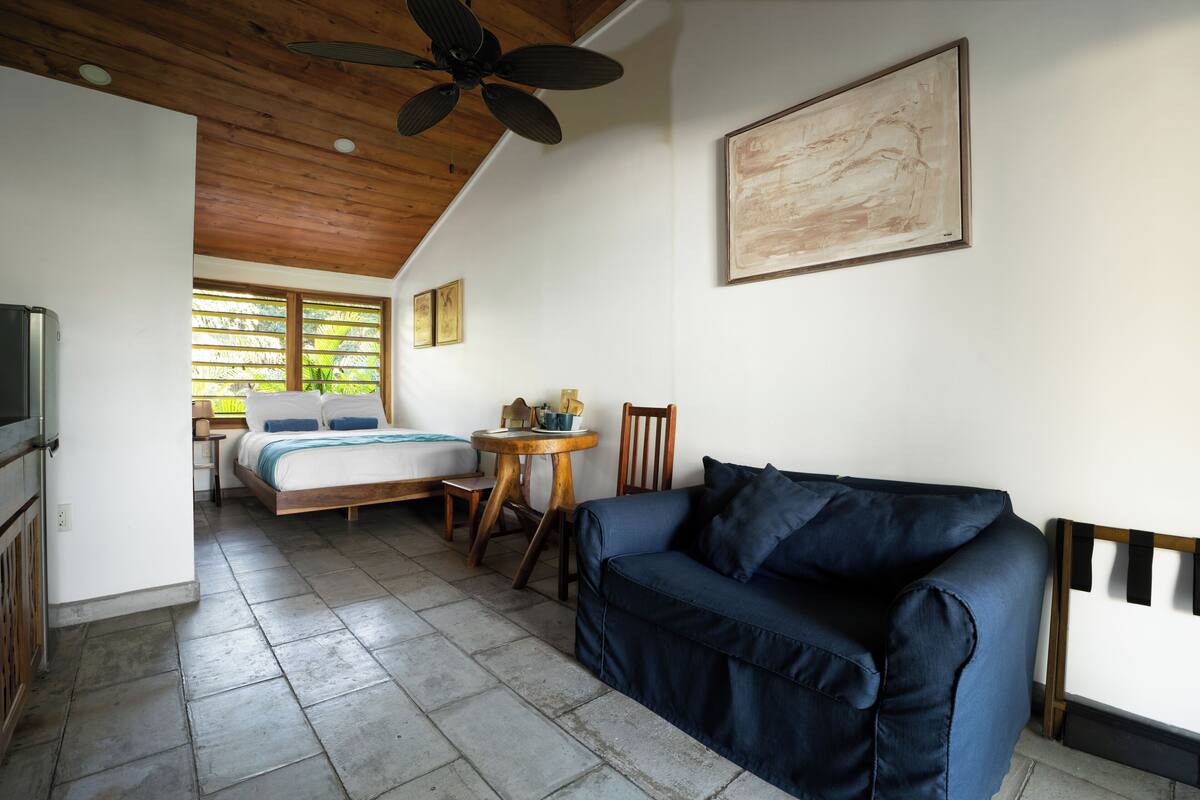 A room with a bed, a blue sofa, a wooden ceiling fan, a painting, and a window with blinds.